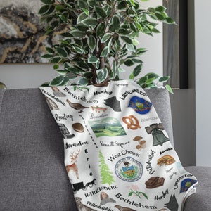 May include: A white throw blanket with a colorful illustration of Pennsylvania landmarks and cities. The blanket features the state seal, pretzels, a Liberty Bell, a cheesesteak, and the names of cities like Philadelphia, Pittsburgh, Hershey, and Lancaster.