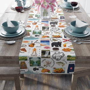 Könnte beinhalten: Ein weißer Tischläufer mit einem sich wiederholenden Muster aus Illustrationen und Text, darunter eine Gitarre, eine Raumfähre und der Text "Dothan". Der Läufer liegt auf einem Holztisch, der für eine Mahlzeit gedeckt ist, mit Tellern, Schalen und Weingläsern.