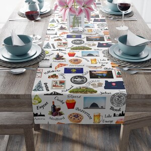 May include: A white table runner with a repeating pattern of Oregon state landmarks and city names. The runner is decorated with images of food, landmarks, and state seals. The table is set with plates, bowls, wine glasses, and flowers.