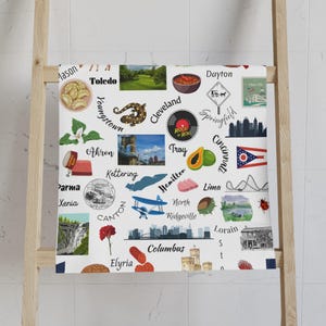 Könnte beinhalten: Weißes Handtuch mit einer Collage von Illustrationen zum Thema Ohio und Städtenamen. Enthält Bilder von Essen, Sehenswürdigkeiten und einer Schallplatte. Städte wie Cleveland, Columbus und Cincinnati sind abgebildet.