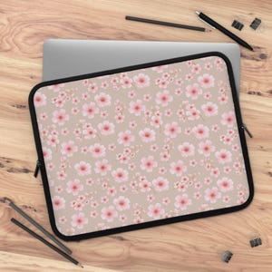 May include: A laptop sleeve featuring a beige background with a repeating pattern of pink cherry blossoms. The sleeve has a black border and a zipper closure. The laptop sleeve is on a wooden surface.
