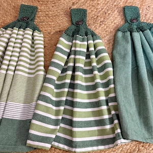 May include: Three hanging kitchen towels. Two are green and white striped, and one is solid teal. Each towel has a green crocheted top with a button closure. The towels are made of a soft fabric.