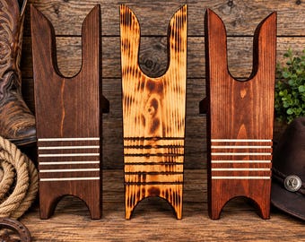 May include: Three wooden boot jacks standing upright. The two outer jacks are dark brown with five horizontal light-coloured stripes. The centre jack features a scorched, flame-treated wood grain pattern with five horizontal grooves across the lower section.