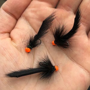 May include: Three black fishing flies with orange beads. Each fly has a black feathery body and a curved hook. The flies are held in a hand, showcasing their details and craftsmanship. These flies are designed for fly fishing.