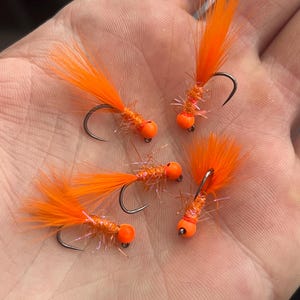 May include: Close-up of four orange fishing flies. Each fly features a black hook, an orange bead, and a vibrant orange feather tail. The flies are designed for angling.