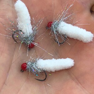 May include: Three white fishing flies with red heads and silver tinsel accents. The flies have a fluffy, textured appearance and are designed with curved black hooks. The flies are designed for fly fishing.