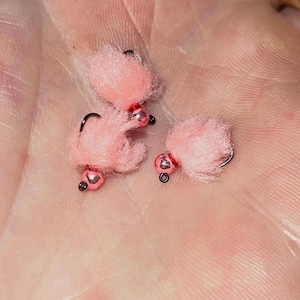 May include: Close-up of three pink fishing flies. Each fly features a fluffy pink body, a small, metallic bead, and a curved black hook. The flies are designed for angling and are likely used to attract fish.