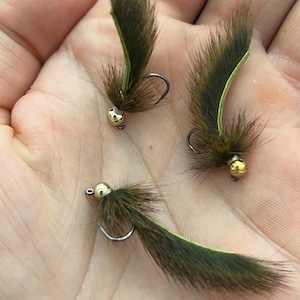 May include: Three fishing flies with dark brown fur bodies, gold-colored heads, and curved hooks. Each fly has a green accent on the tail. The flies are displayed against a light background.