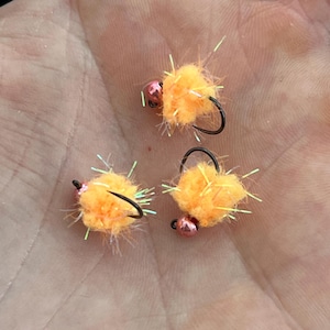 May include: Three orange fishing flies with black hooks and pink beads. The flies have a fluffy, textured appearance with iridescent accents, likely designed to attract fish. The flies are small, and are designed for fly fishing.