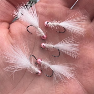 May include: Four white feather fishing flies with pink beads and black hooks are displayed. The flies have a fluffy, feathery appearance, and the pink beads add a pop of color. These flies are designed for fly fishing.