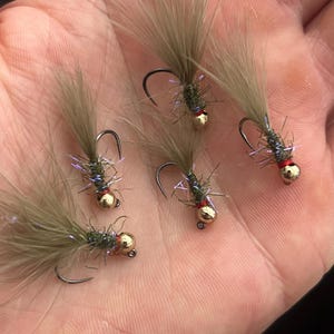 May include: Five fishing flies with gold-colored heads, black hooks, and olive-green bodies. Each fly has a tuft of light brown, feathery material extending from the top. Red accents are visible near the hook.