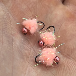 May include: Three handmade fishing flies. Each fly features a fluffy, peach-colored body, a small, pink bead, and a black hook. Iridescent fibers extend from the body, adding visual interest. These flies are designed for angling.
