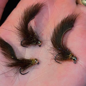 May include: Three brown fishing flies with black hooks and gold-colored beads. The flies have a fluffy, fur-like appearance, with strands of iridescent material. The flies are designed for fly fishing.