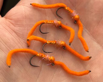 May include: Four bright orange fishing flies with curved black hooks. The flies have a worm-like appearance, with a fuzzy texture and small, rounded heads. The flies are arranged in a hand, suggesting they are ready for use in fly fishing.