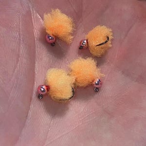 May include: Four orange fishing flies with pink beads and black hooks are displayed. The flies have a fluffy texture and are designed to mimic bait. The flies are arranged on a light pink surface.