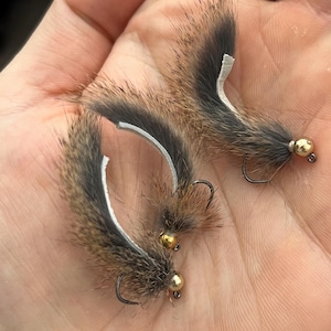 May include: Three handmade fishing flies. Each fly features a brown and gray fur body, a curved white accent, a gold-colored bead head, and a black hook. The flies are designed for fly fishing.