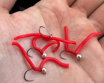 May include: Close-up of several red fishing lures with black hooks and silver beads. The lures are arranged in a hand, showcasing their design and color. These are likely used for catching fish.