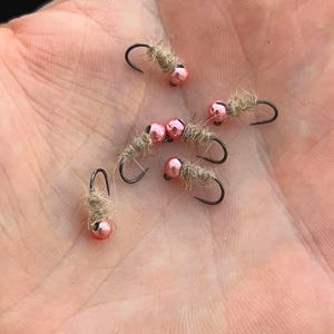 May include: Close-up of several fishing flies. Each fly features a small, pink bead head and a body wrapped in a light brown material. The hooks are black and curved. The flies are arranged on a light-colored surface.