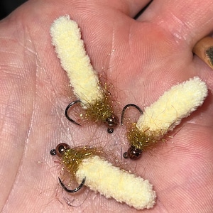 May include: Three handmade fishing flies. Each fly features a cream-colored, fuzzy body, a dark hook, and a brown and gold head. The flies are designed for fly fishing and are shown in a person's hand.