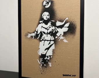 May include: Framed artwork featuring a stencil of a classical figure with outstretched arms, rendered in white against a tan background. The artwork is framed in a black frame, with the artist's name, BANKSY, in black at the bottom right.
