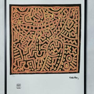 May include: A framed print featuring a vibrant abstract design in black, orange, and yellow. The artwork showcases a complex pattern of interconnected lines and shapes, with the artist's signature in the lower right corner. The print is set against a white background.