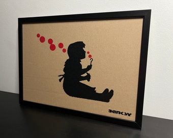 May include: A black and white framed print of a child blowing bubbles. The child is silhouetted against a tan background. The bubbles are red and are floating above the child's head. The print is signed "Banksy" in the bottom right corner.