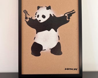 May include: Framed artwork featuring a black and white panda with arms outstretched, holding two guns. The panda is stenciled on a tan background, with the artist's signature "BANKSY" in the lower right corner. The frame is black.