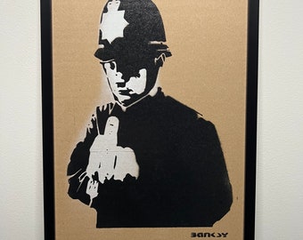 May include: A black and white stencil art print of a police officer giving the middle finger. The print is framed in black and has the artist's signature "Banksy" in white at the bottom.