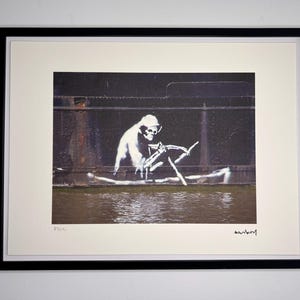 May include: Framed print depicting a white skeleton figure rowing a boat, set against a dark, industrial backdrop. The artwork is signed by Banksy. The print is in a black frame with a white mount.