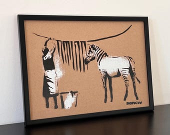 May include: Black and white stencil art print of a woman washing a zebra with a bucket of water. The art is framed in a black frame and signed "Banksy" in the bottom right corner.