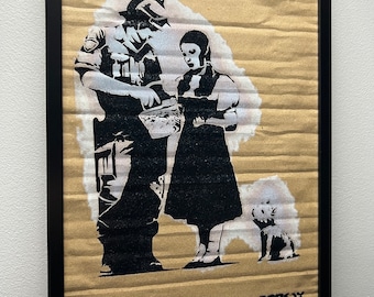 May include: Black and white stencil art print on cardboard featuring a police officer giving a bucket to a girl in a dress. A small white dog is standing behind the girl. The artwork is signed "Banksy" in the bottom right corner.