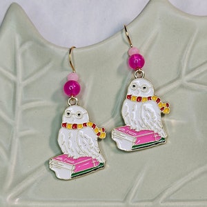 May include: A pair of gold earrings featuring a white owl with pink and yellow accents perched on a stack of pink books. The owl is wearing glasses and a scarf. The earrings are accented with pink beads.