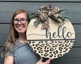 May include: A round wooden door hanger with a leopard print design and the word "hello" in black script. A brown and black leopard print bow is attached to the top of the door hanger.