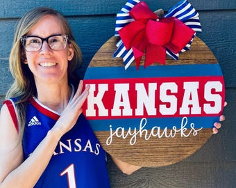 May include: A wooden round door hanger with a red, white, and blue striped design. The words "KANSAS jayhawks" are written in white on the door hanger. A red and white striped bow with a red center is attached to the top of the door hanger.