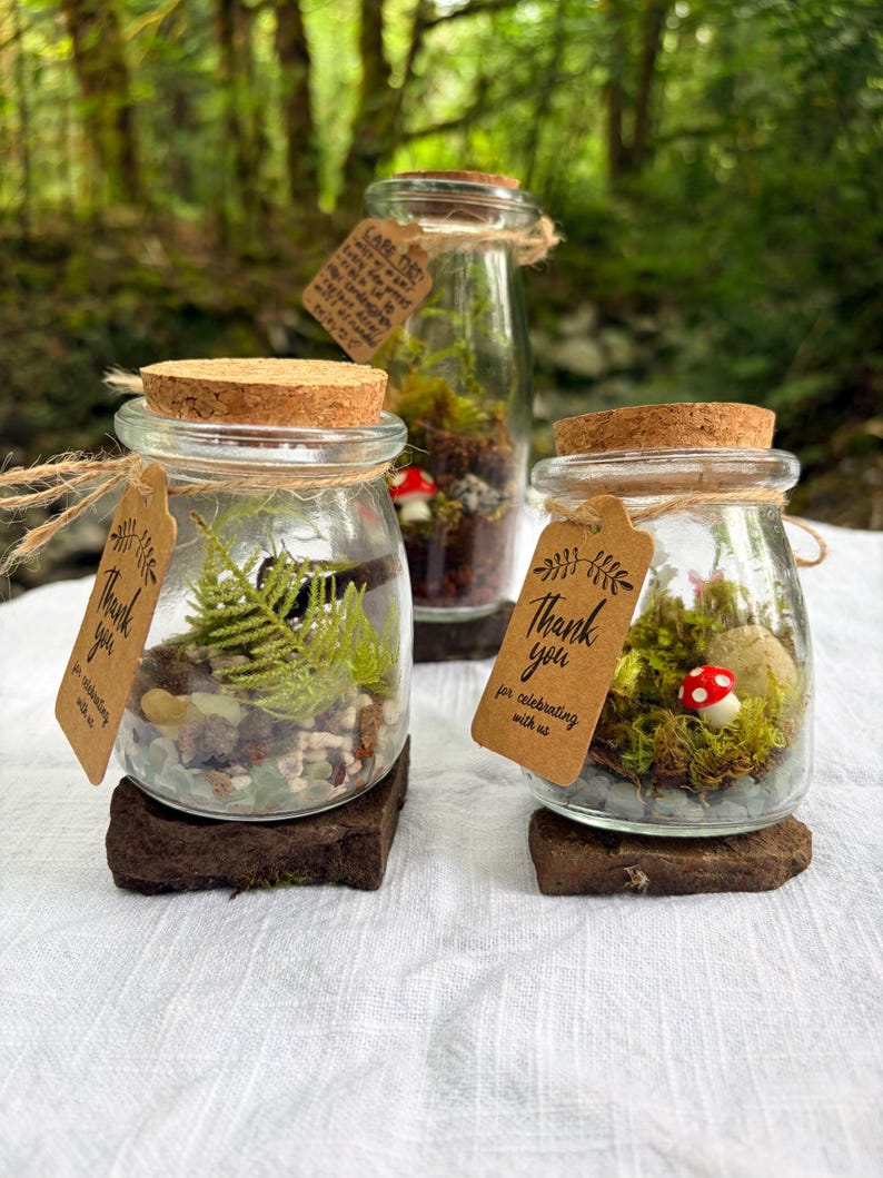 May include: Three glass jars with cork lids, each containing a miniature terrarium with moss, stones, and a small red and white mushroom. Each jar features a brown paper tag tied with twine, printed with the text Thank you for celebrating with us.