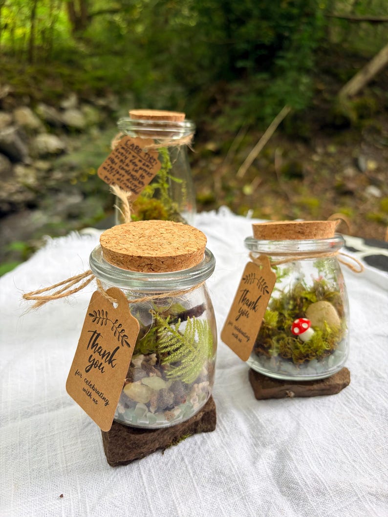 May include: Two small glass jars with cork lids containing miniature moss terrariums with green ferns, small stones, and a red mushroom figurine. Each jar features a brown paper tag tied with twine that says Thank you for celebrating with us.