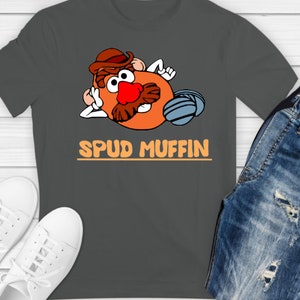 Op de afbeelding: Een donkergrijs T-shirt met een cartoon afbeelding van Mr. Potato Head met een hoed en zonnebril. De tekst "SPUD MUFFIN" is in oranje onder de afbeelding gedrukt.