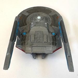 Puede incluir: Una maqueta gris de nave espacial con detalles en azul y rojo. El modelo tiene dos alas extendidas con detalles azules y un cuerpo central con detalles intrincados. La parte superior presenta un dise&ntilde;o circular y una luz central.