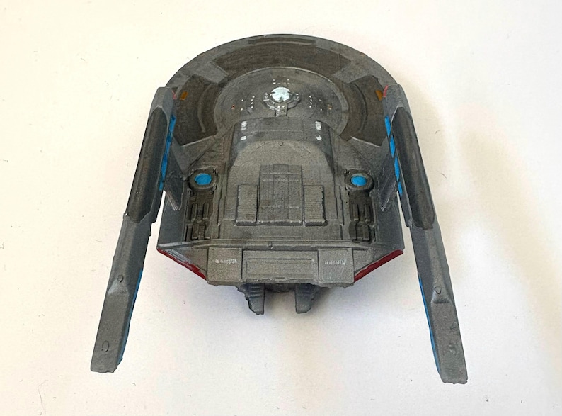 Puede incluir: Una maqueta gris de nave espacial con detalles en azul y rojo. El modelo tiene dos alas extendidas con detalles azules y un cuerpo central con detalles intrincados. La parte superior presenta un dise&ntilde;o circular y una luz central.