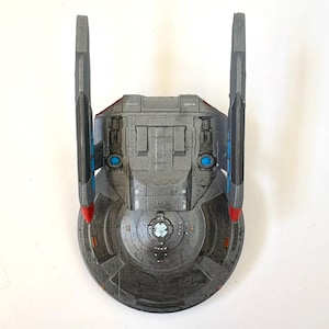 Puede incluir: Modelo gris de una nave espacial con dos aletas verticales y una base circular. La nave tiene detalles en azul y rojo, con un dise&ntilde;o detallado en el cuerpo principal. El modelo parece ser una r&eacute;plica de una nave espacial de ciencia ficci&oacute;n.