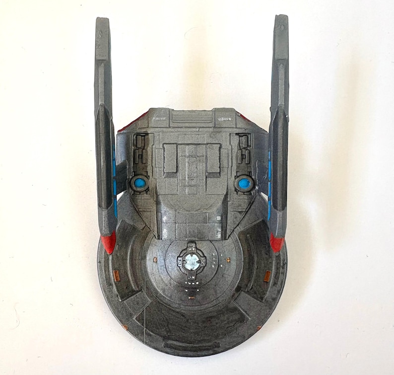 Puede incluir: Modelo gris de una nave espacial con dos aletas verticales y una base circular. La nave tiene detalles en azul y rojo, con un dise&ntilde;o detallado en el cuerpo principal. El modelo parece ser una r&eacute;plica de una nave espacial de ciencia ficci&oacute;n.