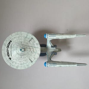 Könnte beinhalten: Ein Modell des Raumschiffs USS Enterprise NCC-1701-A. Das Modell ist hauptsächlich weiß und grau mit blauen Akzenten auf den Gondeln. Die Bezeichnung des Schiffs steht in schwarzer Schrift auf der Untertasse. Sammlerobjekt.