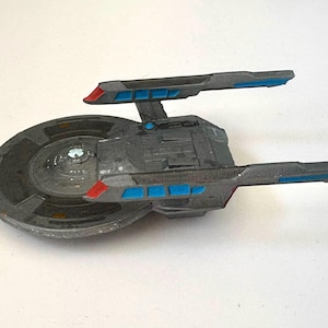 Puede incluir: Modelo gris de una nave espacial con detalles azules y rojos. La nave tiene un cuerpo principal circular y dos largas extensiones rectangulares. Las extensiones tienen detalles rectangulares azules y puntas rojas. El modelo est&aacute; sobre una superficie blanca.