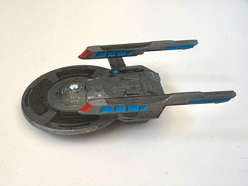 Puede incluir: Modelo gris de una nave espacial con detalles azules y rojos. La nave tiene un cuerpo principal circular y dos largas extensiones rectangulares. Las extensiones tienen detalles rectangulares azules y puntas rojas. El modelo est&aacute; sobre una superficie blanca.