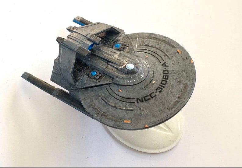 Puede incluir: Maqueta gris de una nave espacial con un casco circular con el texto "NCC-31080-A". El modelo tiene detalles azules y cobrizos y est&aacute; montado sobre un soporte blanco. Un art&iacute;culo de colecci&oacute;n.