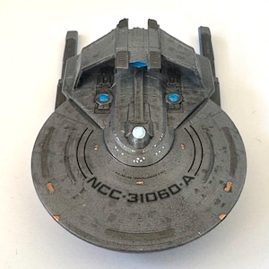 Puede incluir: Maqueta gris de nave espacial con el texto "NCC-31060-A" impreso en la parte delantera. El modelo tiene una secci&oacute;n frontal circular y una secci&oacute;n superior rectangular con detalles azules y blancos. R&eacute;plica de una nave espacial.