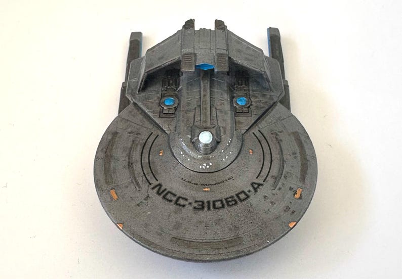 Puede incluir: Maqueta gris de nave espacial con el texto "NCC-31060-A" impreso en la parte delantera. El modelo tiene una secci&oacute;n frontal circular y una secci&oacute;n superior rectangular con detalles azules y blancos. R&eacute;plica de una nave espacial.