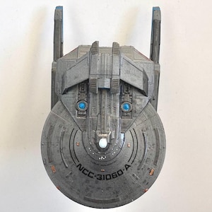 Puede incluir: Maqueta gris de nave espacial con detalles azules y naranjas. La nave tiene una secci&oacute;n frontal circular con el texto "NCC-31060-A". Dos luces azules son visibles en la parte delantera, y dos estructuras verticales se extienden hacia arriba.