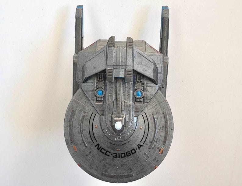Puede incluir: Maqueta gris de nave espacial con detalles azules y naranjas. La nave tiene una secci&oacute;n frontal circular con el texto "NCC-31060-A". Dos luces azules son visibles en la parte delantera, y dos estructuras verticales se extienden hacia arriba.