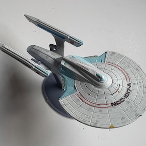 Puede incluir: Maqueta de la nave estelar Enterprise NCC-1701-A, una nave espacial con casco gris, detalles azules y dos góndolas de curvatura. La nave tiene un casco principal circular y está montada sobre una base azul oscuro.